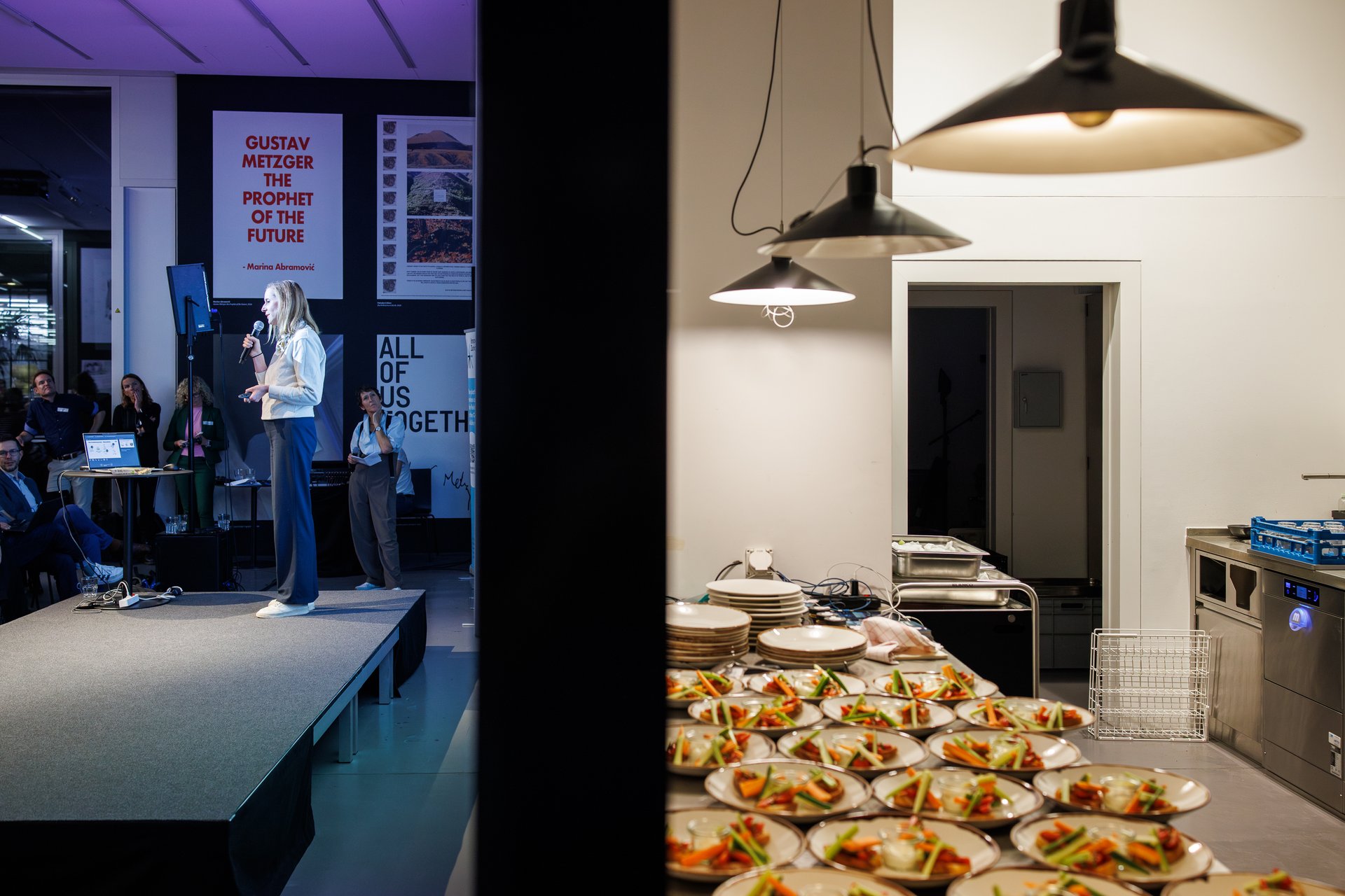 A person stands on a small stage and speaks to an audience. On the left side of the image, spectators can be seen, and on the right side, an adjoining room with many prepared plate portions on a work surface.