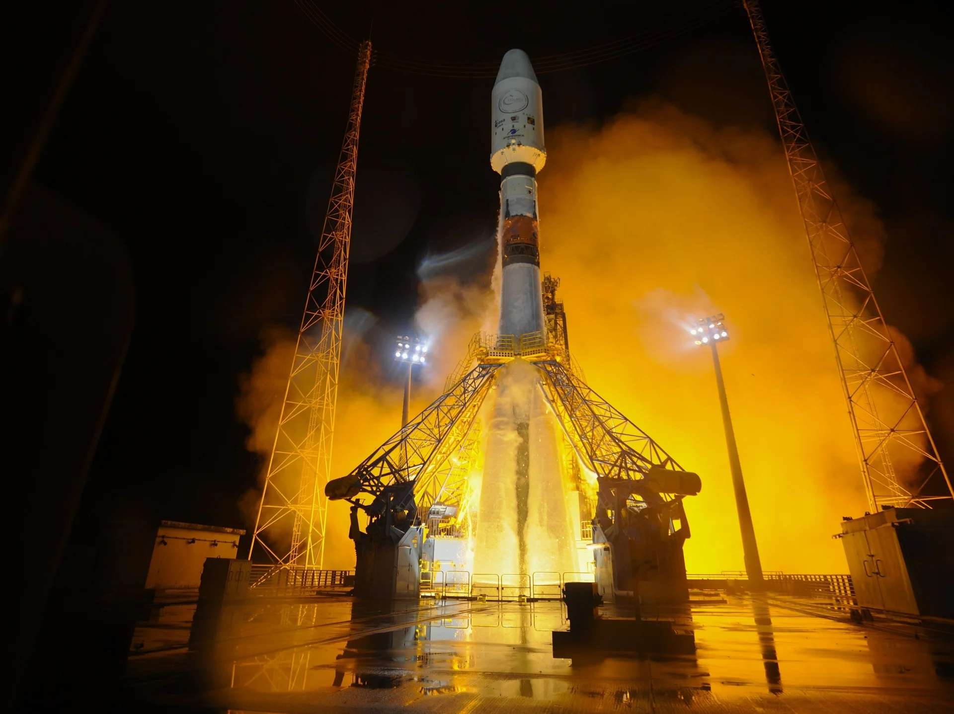 Rakete auf einer beleuchteten Startrampe bei Nacht, umgeben von orangefarbenem Rauch und Dampf.