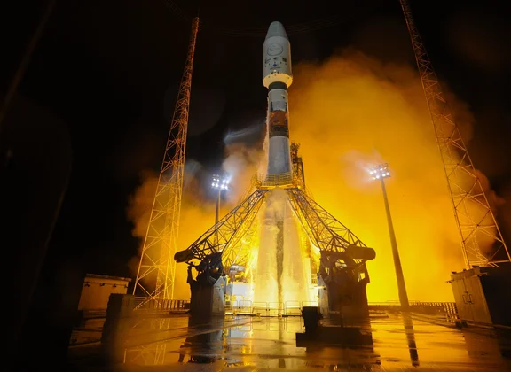 A rocket on a floodlit launch pad at night, surrounded by orange smoke and steam.