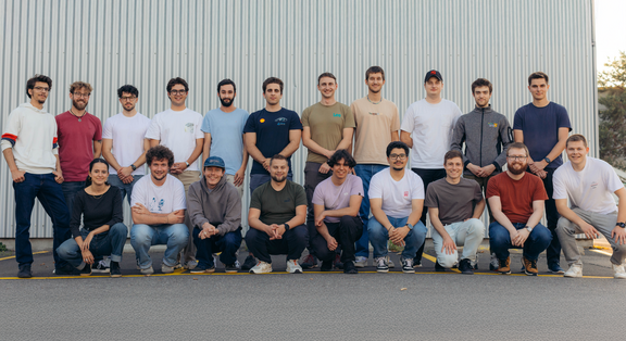 Group photo of the Racing East team: A total of 20 team members are standing and kneeling in two rows in front of a grey metal wall. Most of them are wearing casual clothes such as T-shirts, hoodies and jeans and smiling at the camera. The picture conveys a friendly, motivated team spirit.