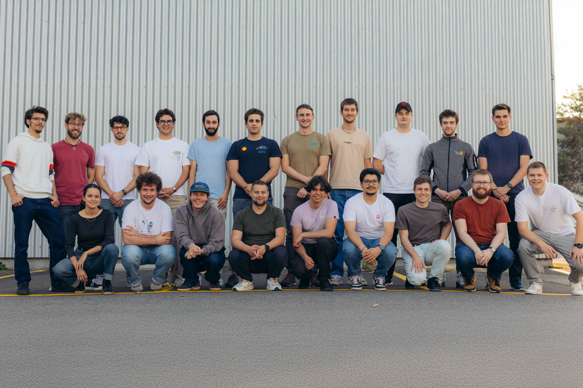 Group photo of the Racing East team: A total of 20 team members are standing and kneeling in two rows in front of a grey metal wall. Most of them are wearing casual clothes such as T-shirts, hoodies and jeans and smiling at the camera. The picture conveys a friendly, motivated team spirit.
