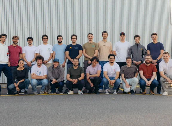 Gruppenfoto des Racing-Ost-Teams: Insgesamt 20 Teammitglieder stehen und knien in zwei Reihen vor einer grauen Metallwand. Die Personen tragen überwiegend lässige Kleidung wie T-Shirts, Hoodies und Jeans und lächeln in die Kamera. Das Bild vermittelt ein freundschaftliches, motiviertes Teamgefühl.