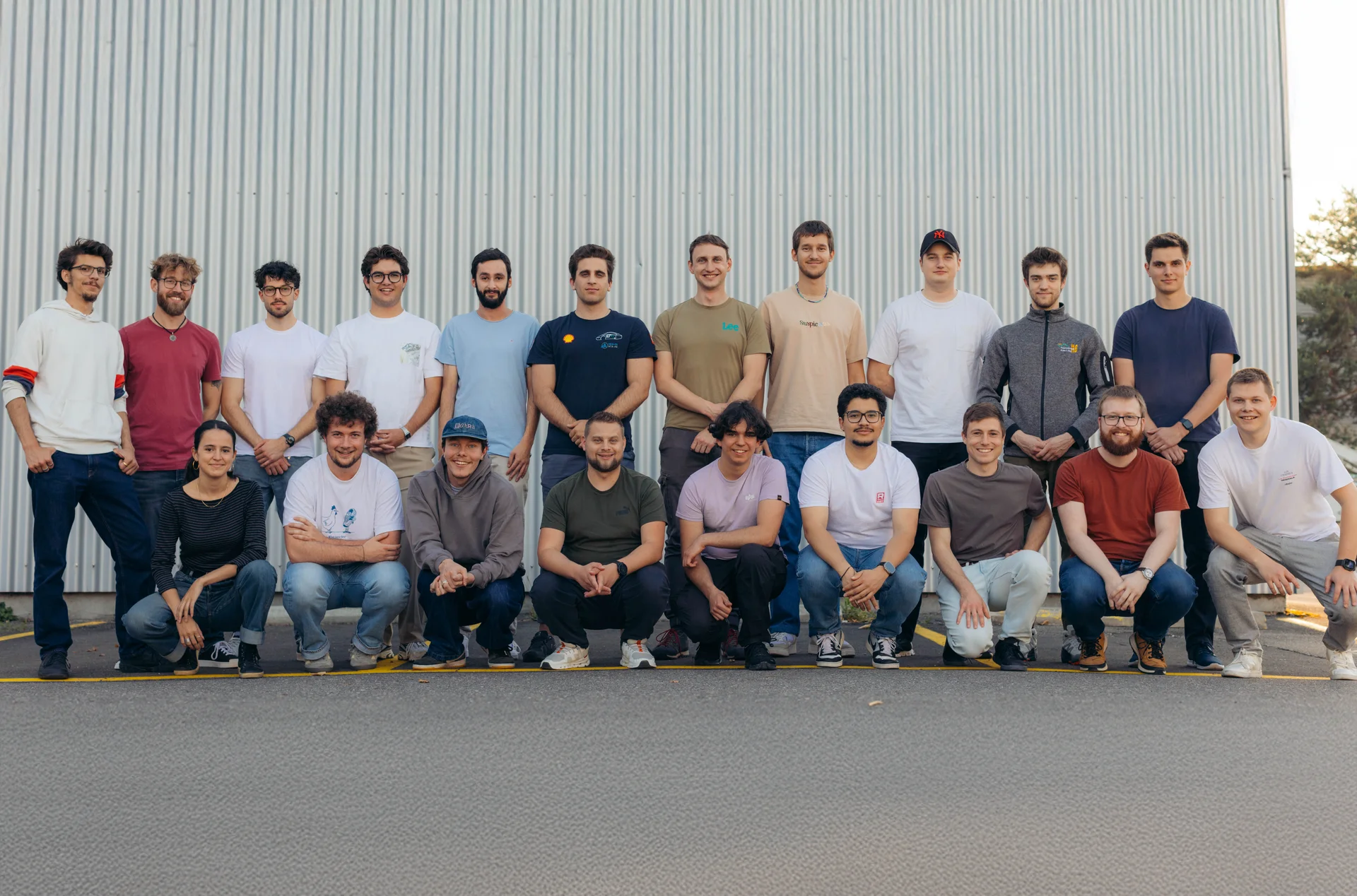 Gruppenfoto des Racing-Ost-Teams: Insgesamt 20 Teammitglieder stehen und knien in zwei Reihen vor einer grauen Metallwand. Die Personen tragen überwiegend lässige Kleidung wie T-Shirts, Hoodies und Jeans und lächeln in die Kamera. Das Bild vermittelt ein freundschaftliches, motiviertes Teamgefühl.