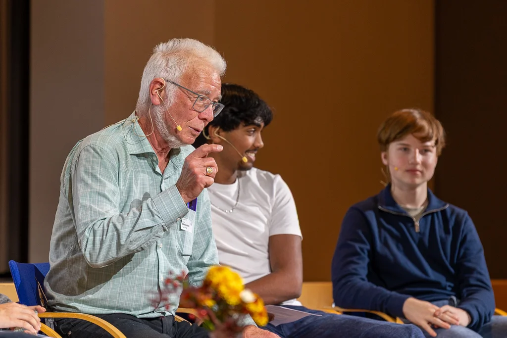 Older man speaks on the podium, two young men on the podium listen intently.