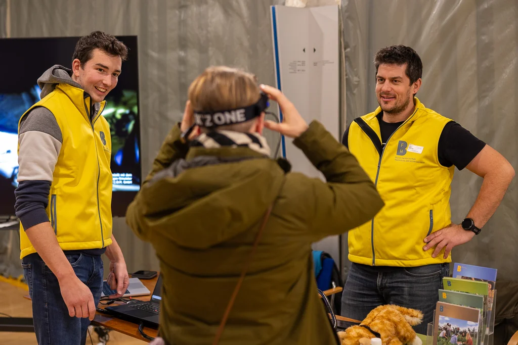 Visitor with VR glasses at a stand with two men in yellow waistcoats.
