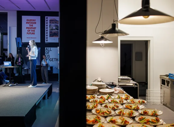A person stands on a small stage and speaks to an audience. On the left side of the image, spectators can be seen, and on the right side, an adjoining room with many prepared plate portions on a work surface.