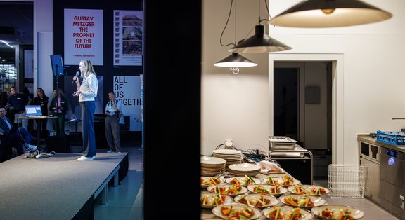 A person stands on a small stage and speaks to an audience. On the left side of the image, spectators can be seen, and on the right side, an adjoining room with many prepared plate portions on a work surface.